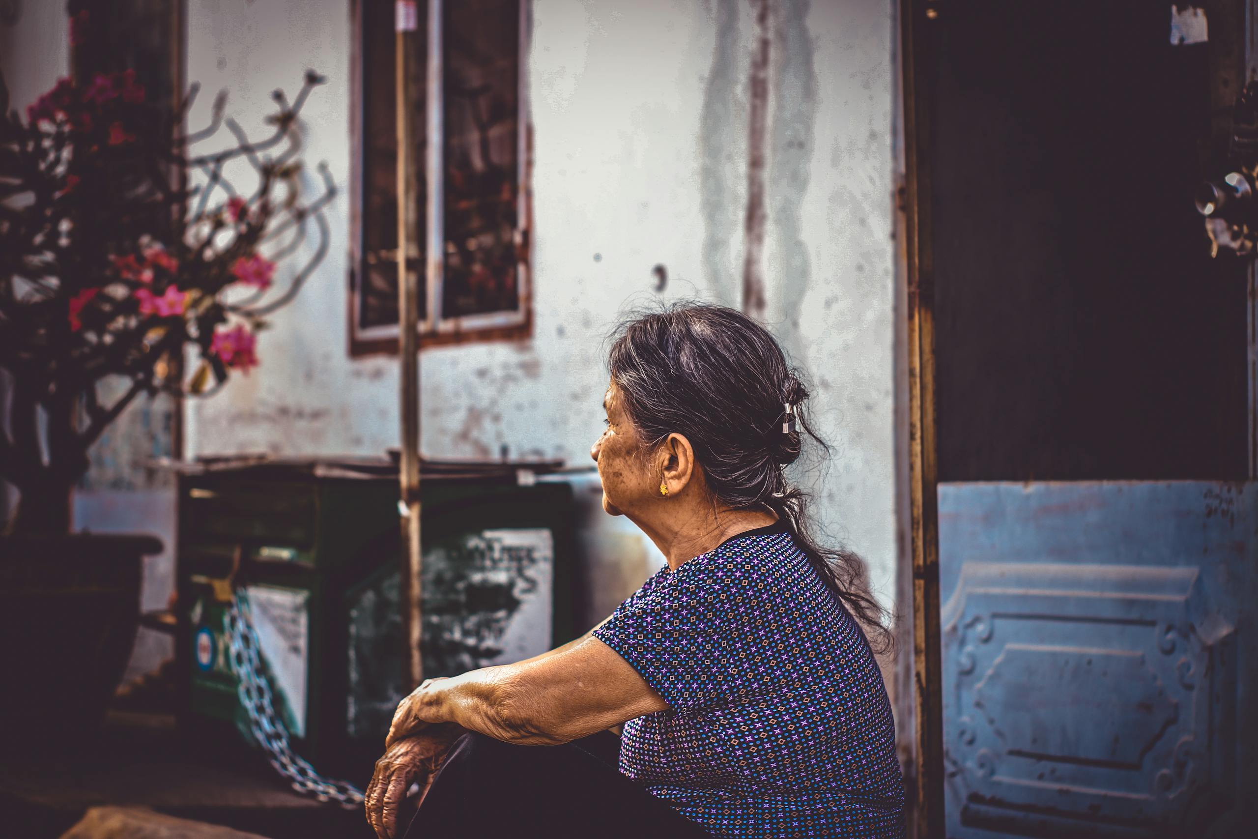 Elderly woman sitting by a doorway, gazing thoughtfully outdoors in a rustic setting.