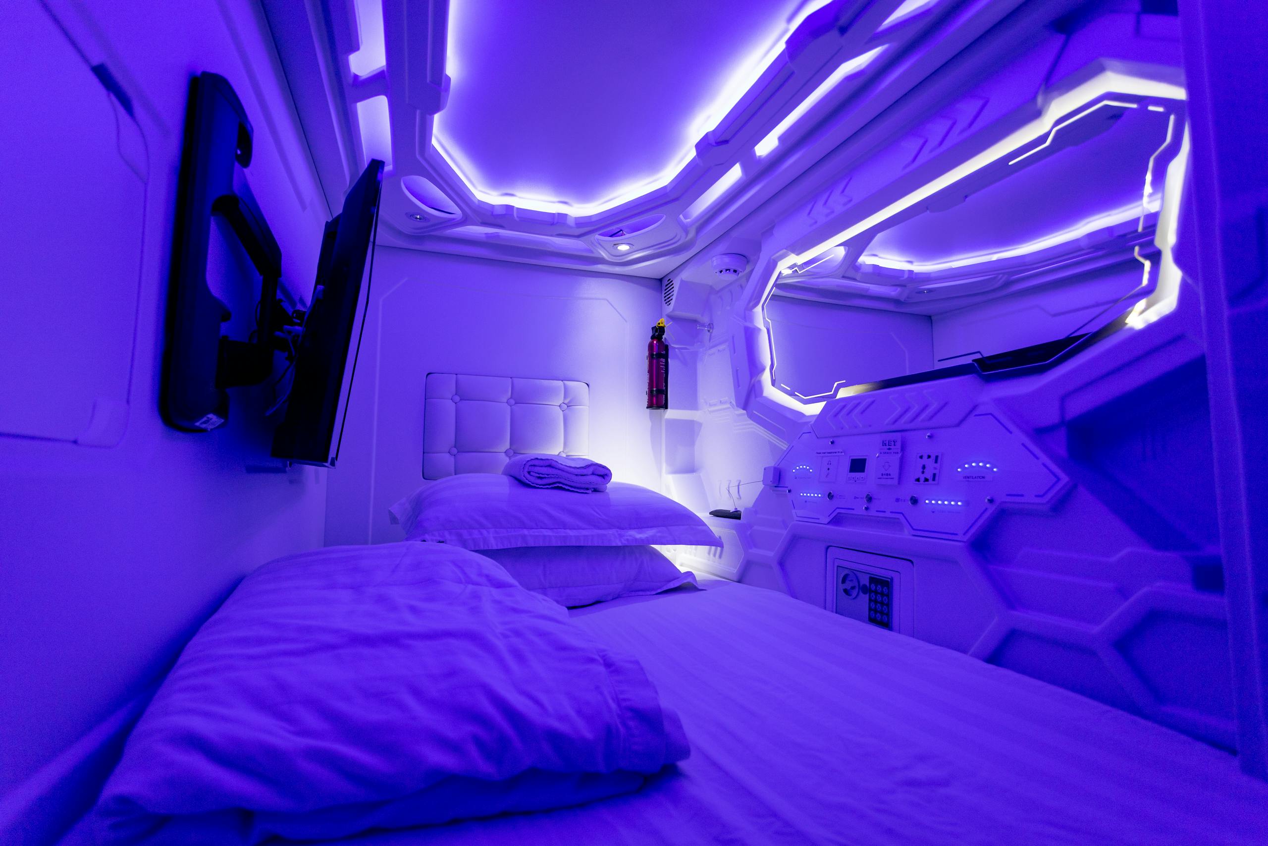 Explore a futuristic capsule hotel room adorned with calming ultraviolet lighting, perfect for a tranquil stay.