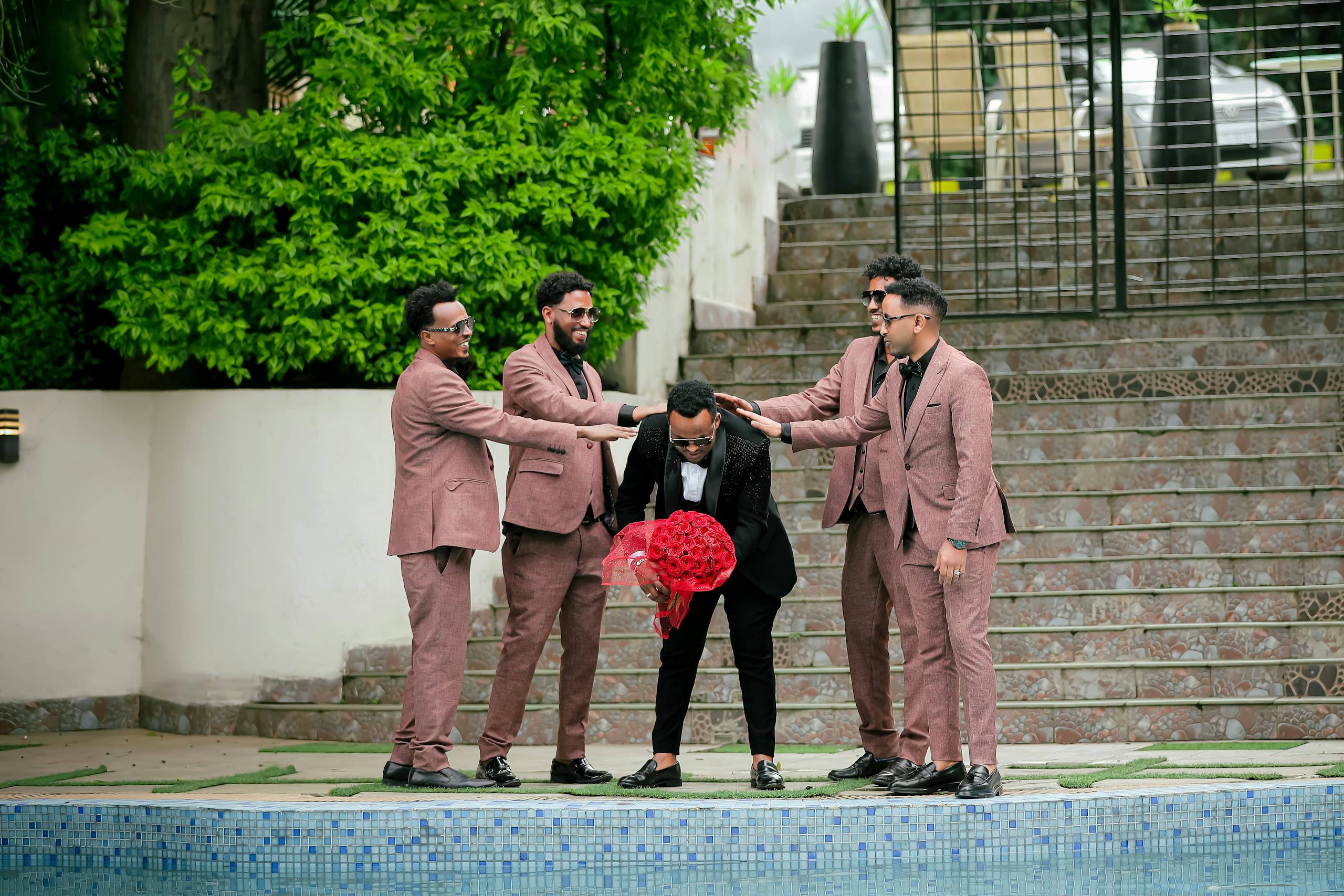 Five groomsmen in suits have fun with the groom holding a red bouquet outdoors by a pool.