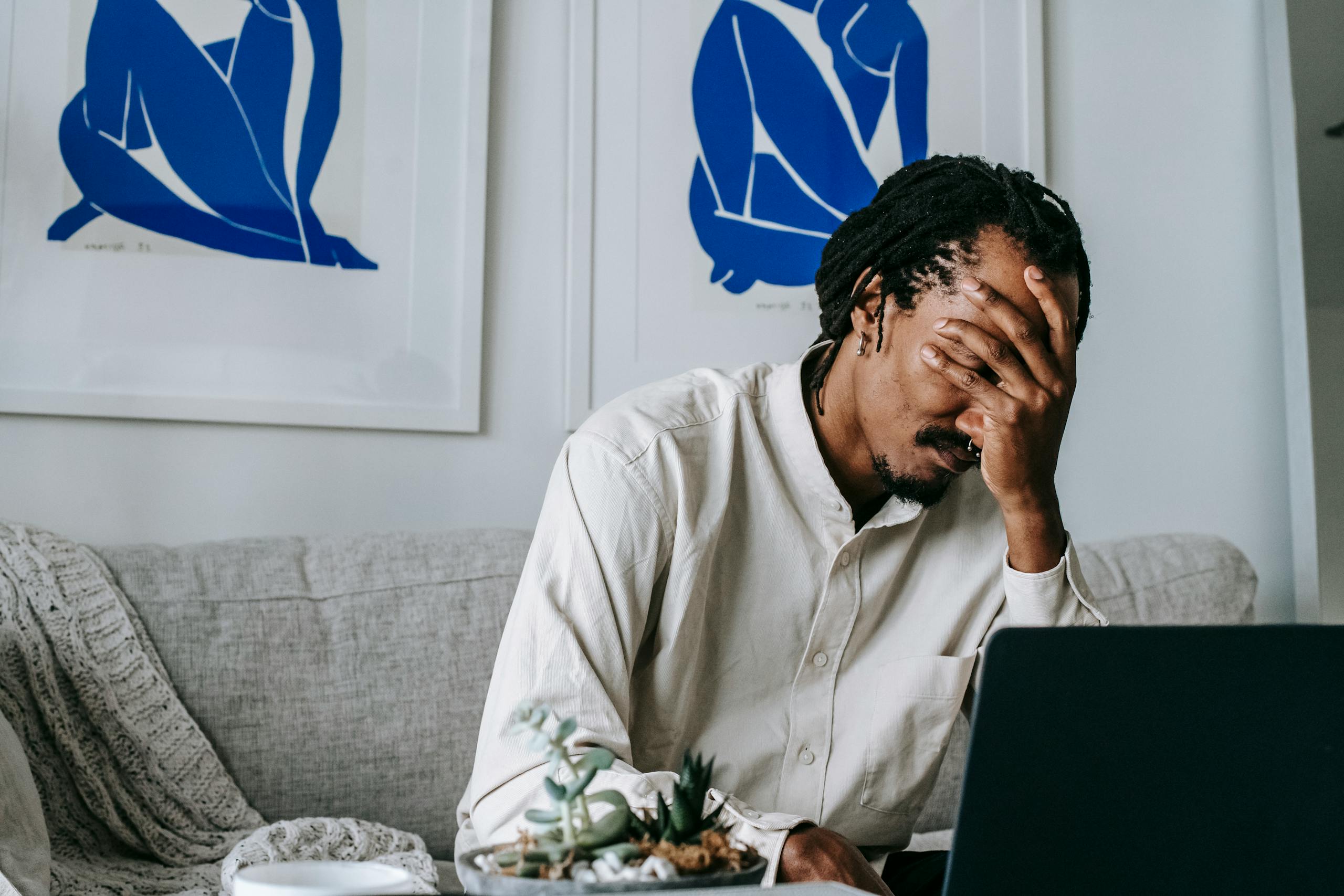 Frustrated young bearded African American male freelancer with dreadlocks in casual shirt working on laptop at home and covering eyes with hand after failure