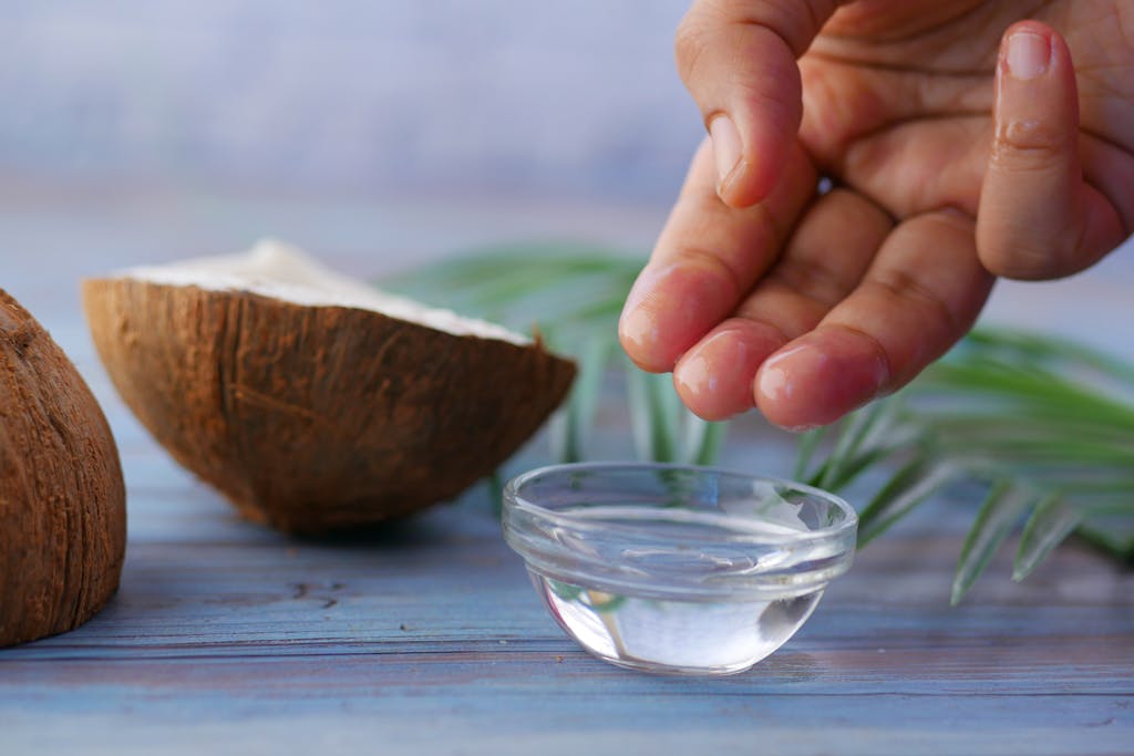Hands dipping into coconut oil bowl beside fresh coconut halves on wooden surface