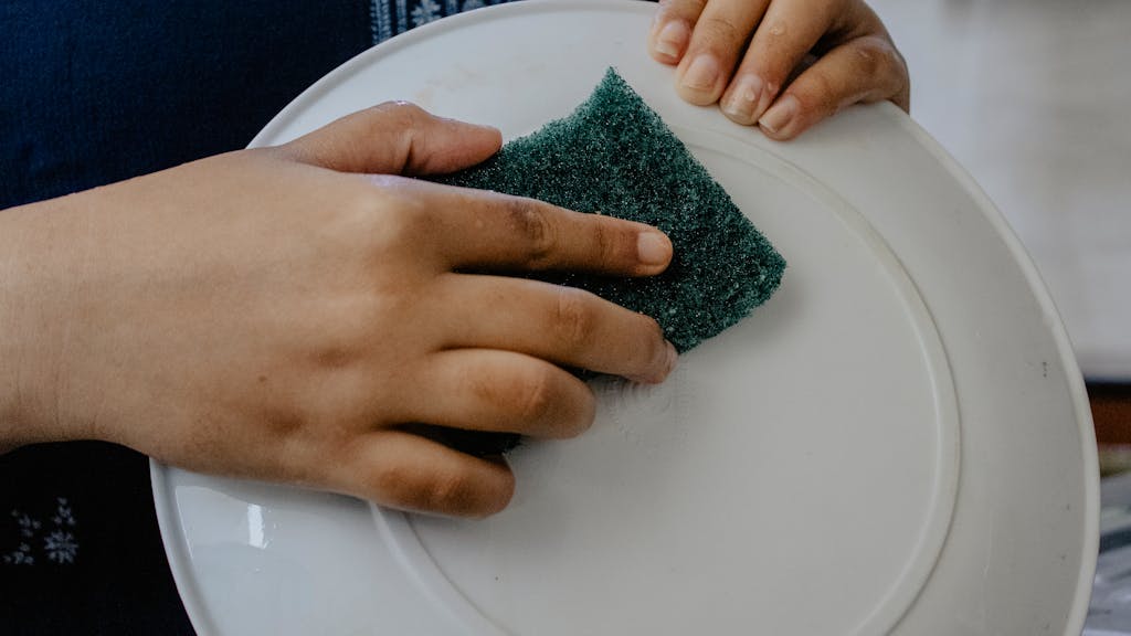 Hands using a green scrub pad to clean a white plate indoors.