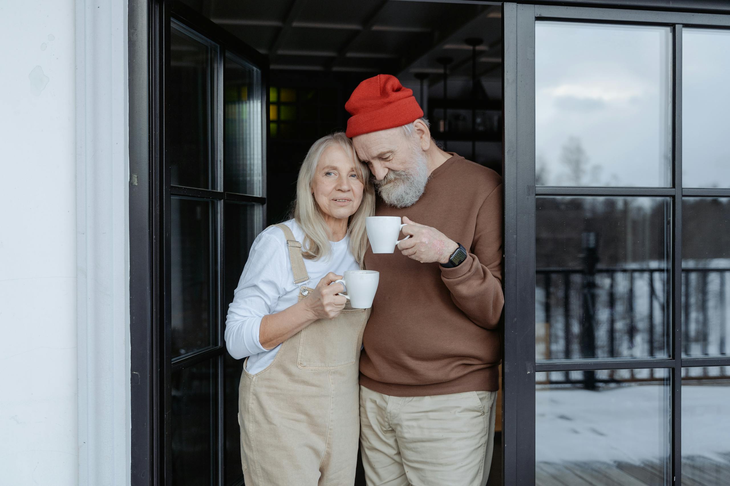 Happy senior couple stands with coffee by the door on a snowy winter day.