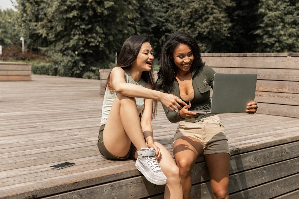 Happy young multiracial female friends in casual clothes laughing while watching funny video on laptop sitting on wooden podium in park