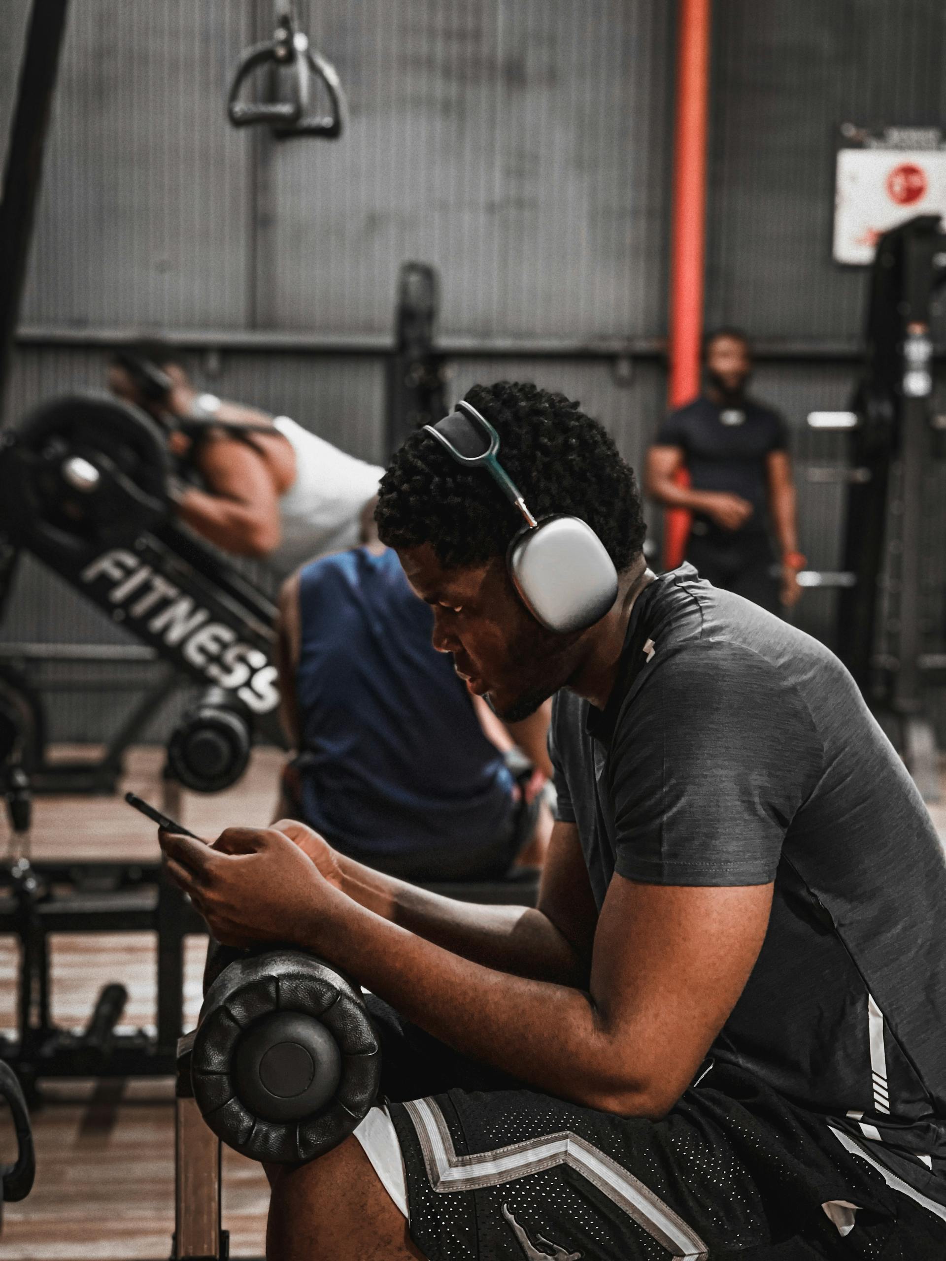 Man sitting in gym listening to music on headphones, using smartphone. Fitness and relaxation.