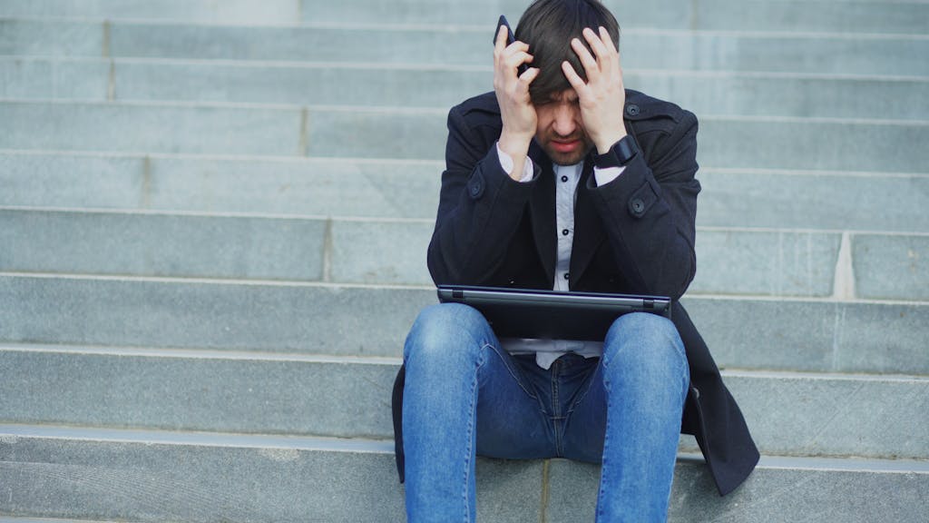 Man sitting on stairs with laptop, looking stressed and frustrated.