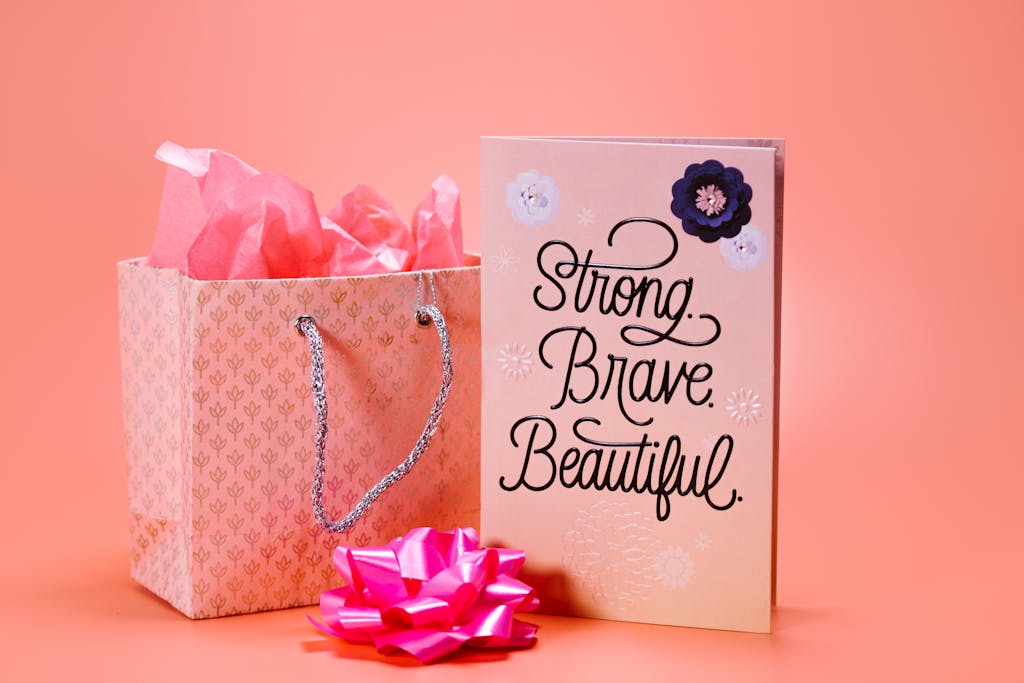 Pink gift bag and card with 'Strong. Brave. Beautiful.' text on a peach background.