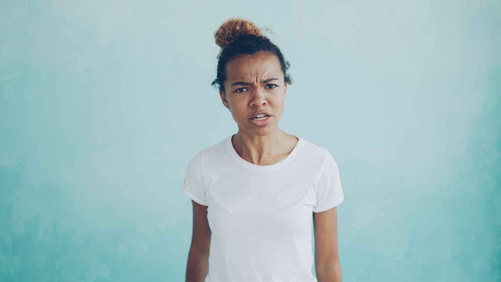 Portrait of a woman in a white t-shirt expressing anger against a blue background.