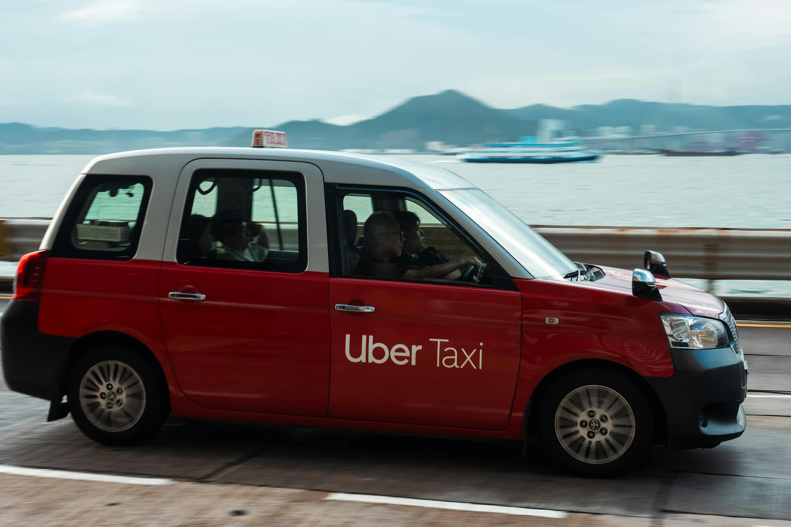 Red Uber taxi driving along a waterfront with a serene view of the sea and distant mountains.