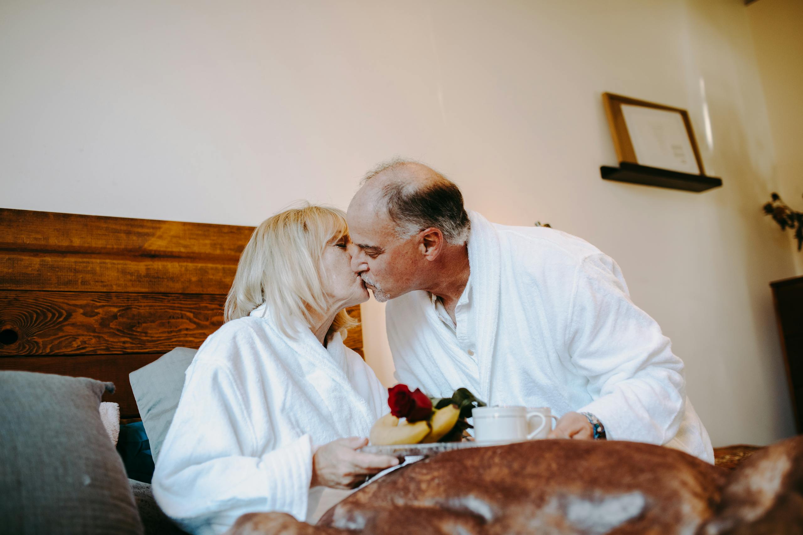 Senior couple kissing with breakfast and roses in bed, showcasing love and intimacy.