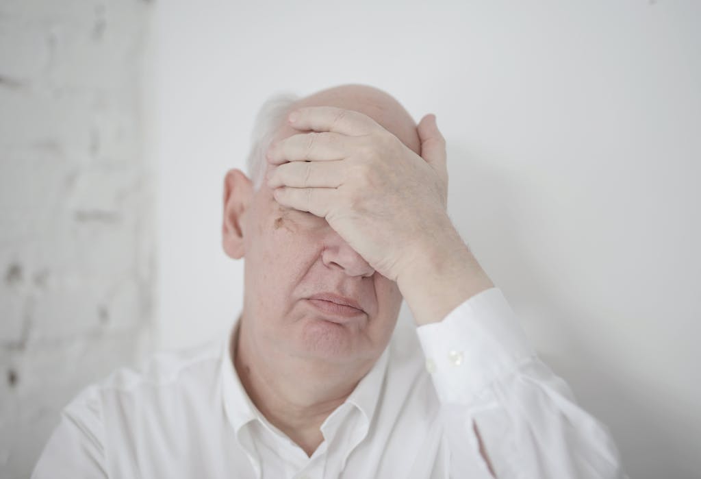 Senior man in white shirt covering his face with hand indoors, expressing thoughtful emotions.