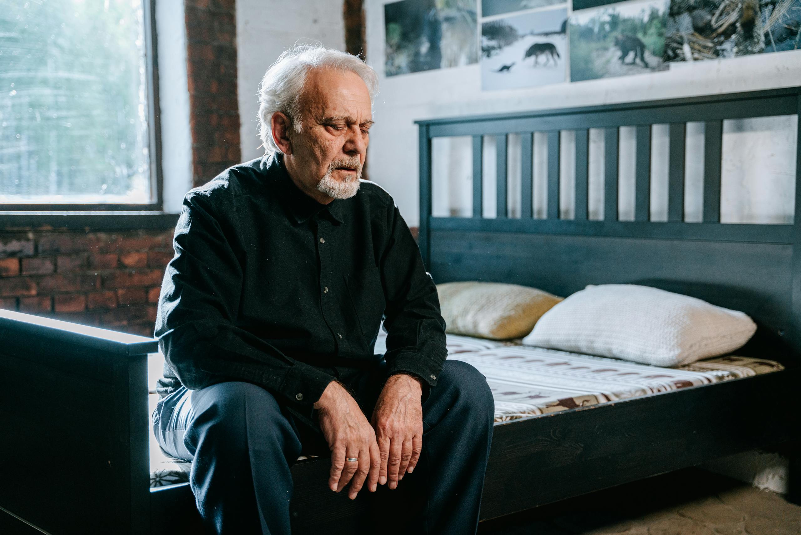 Senior man sitting on bed in pensive mood in a dimly lit bedroom.