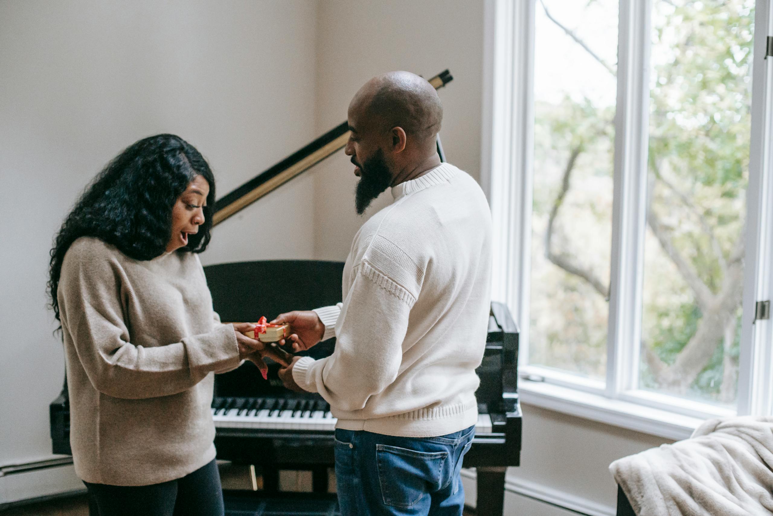 Side view of surprised African American woman receiving present from beaded husband during holiday celebration in room with piano near window