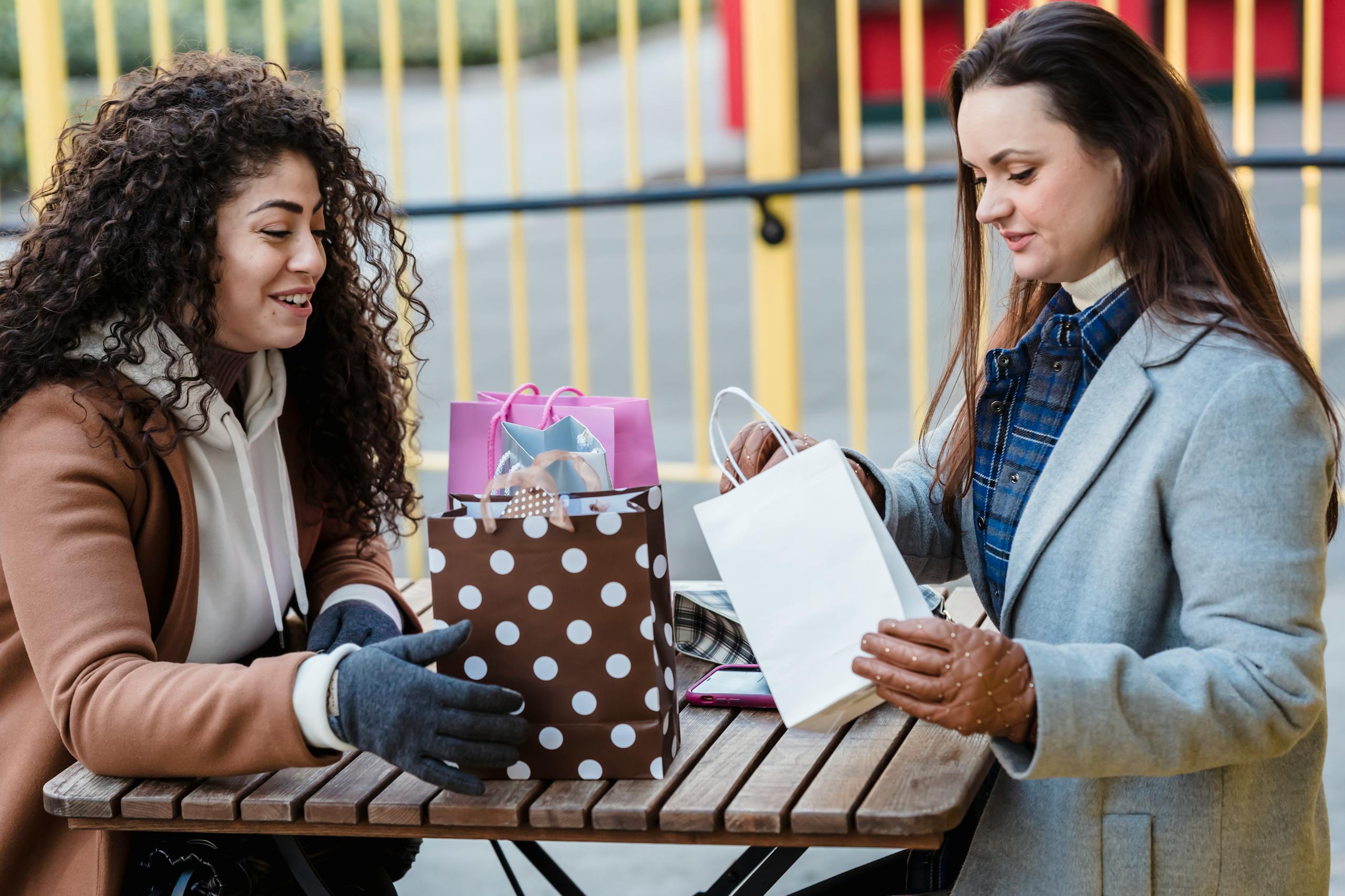 Side view of young female friends in warm coats and gloves sitting at table and checking gift packages in chairs in street near yellow fence in town in daylight