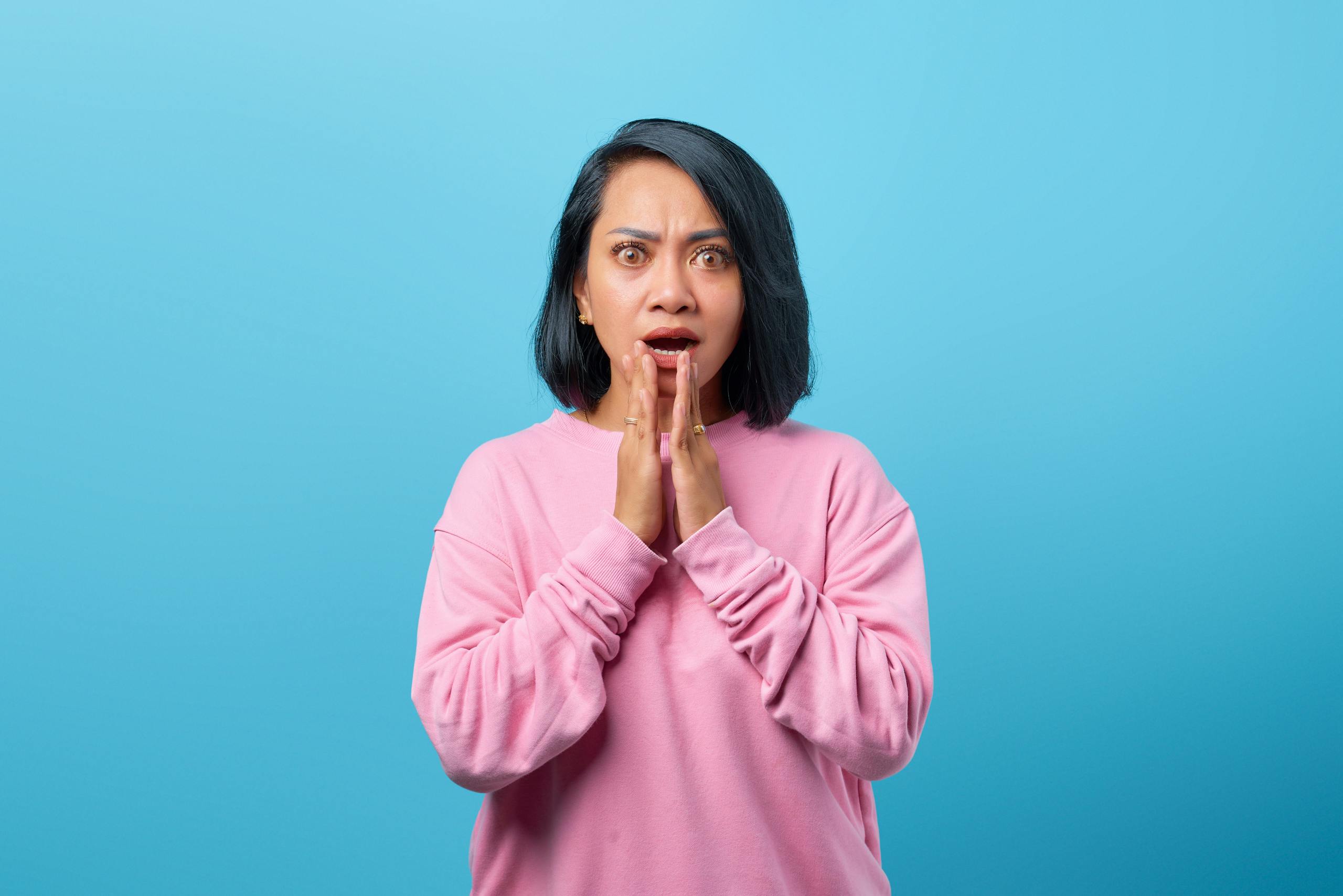 Surprised woman in a pink sweatshirt with hands on chin against a bright blue backdrop, expressing shock.