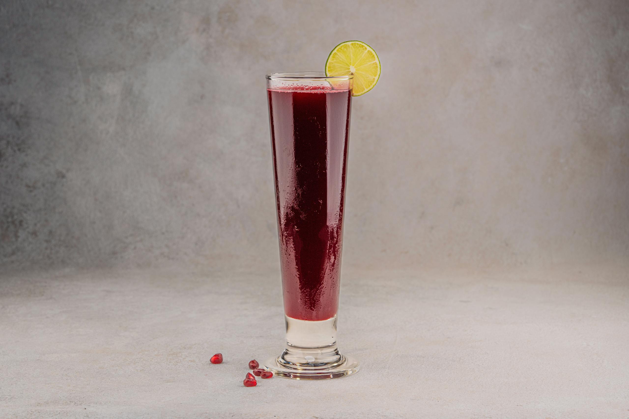 Tall glass of vibrant pomegranate juice with a lemon slice on a neutral background.