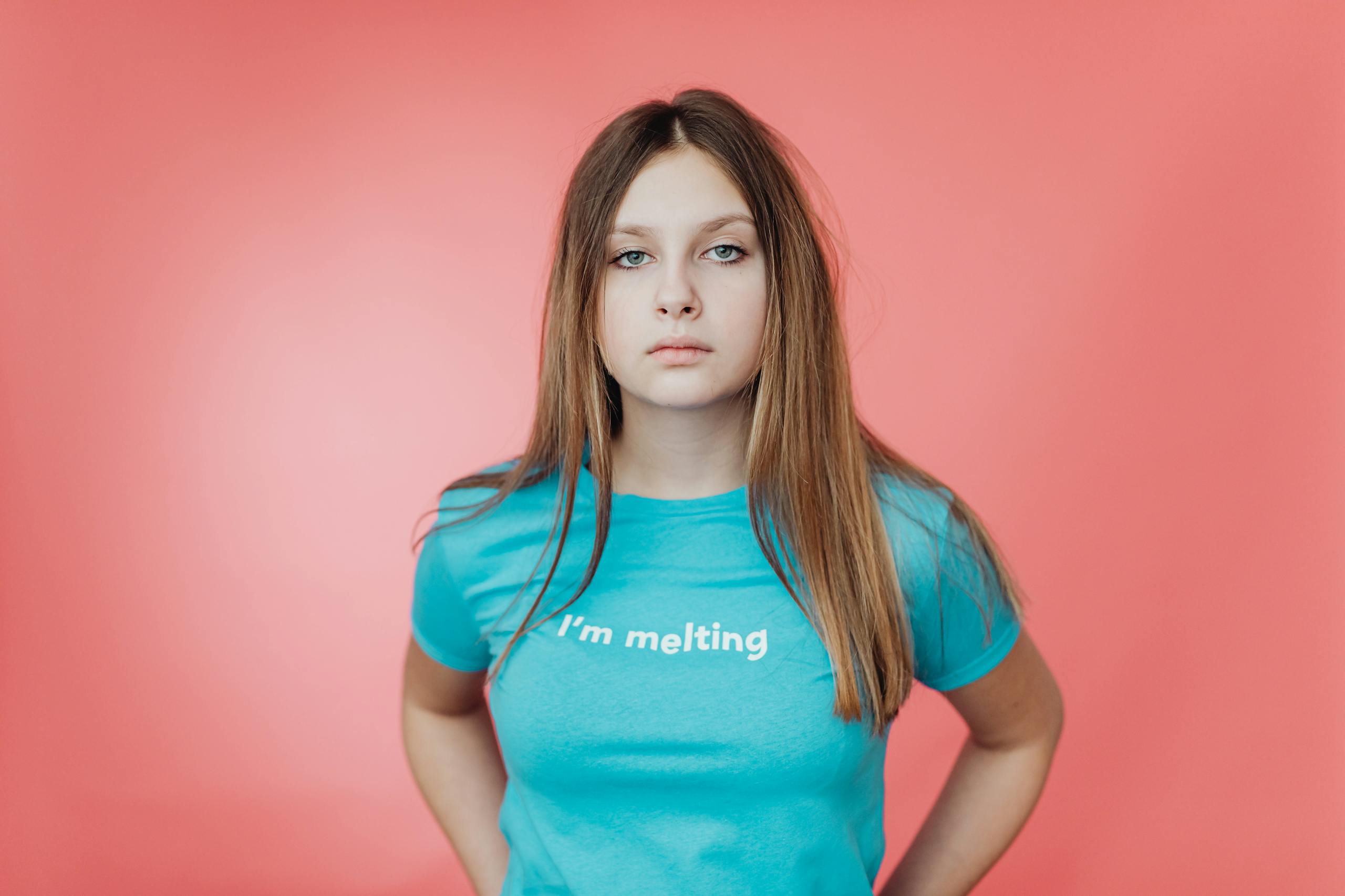 Teen girl wearing a blue shirt with 'I’m melting' text, against a pink backdrop.