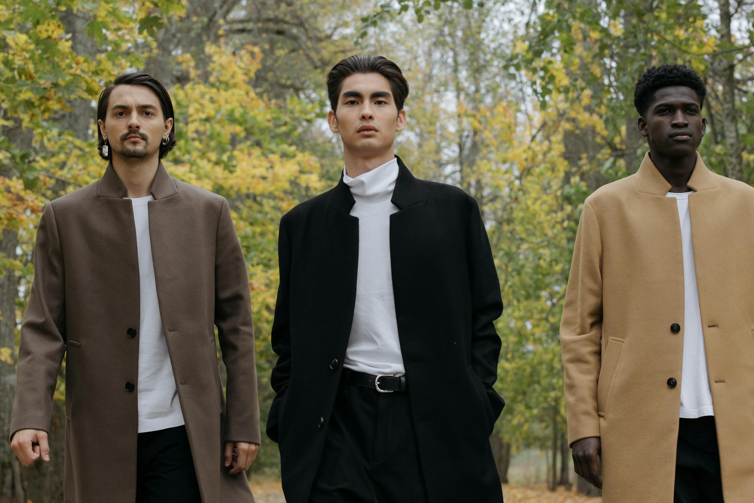 Three men in trendy coats walk confidently through an autumnal park, showcasing contemporary fashion.