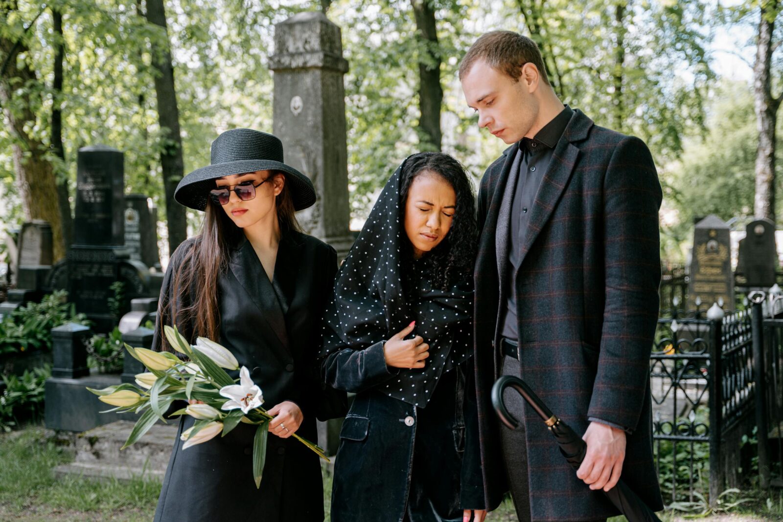 Three mourners in a cemetery, expressing grief amidst tombstones and greenery.