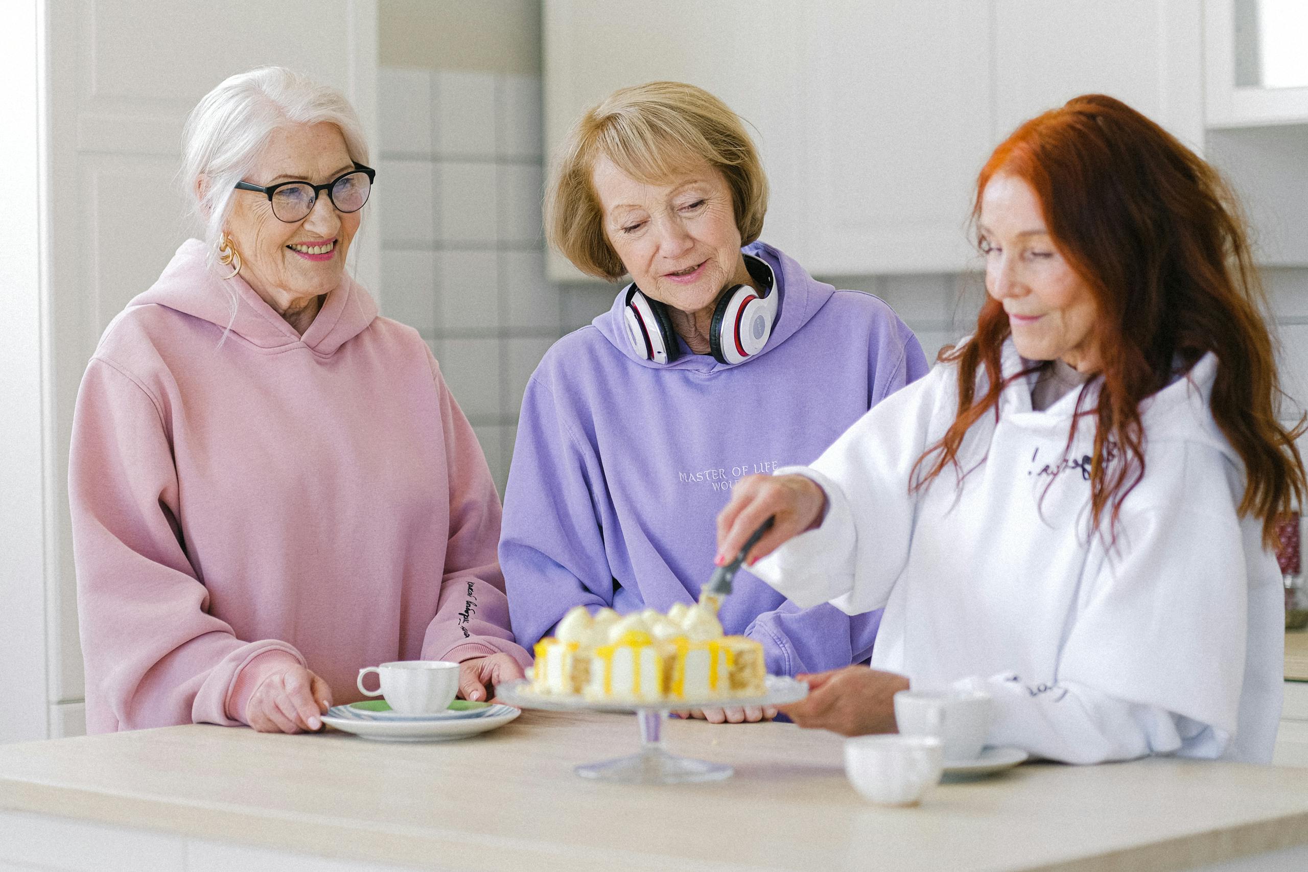 Three senior women happily gathering in a kitchen, enjoying cake and coffee together.