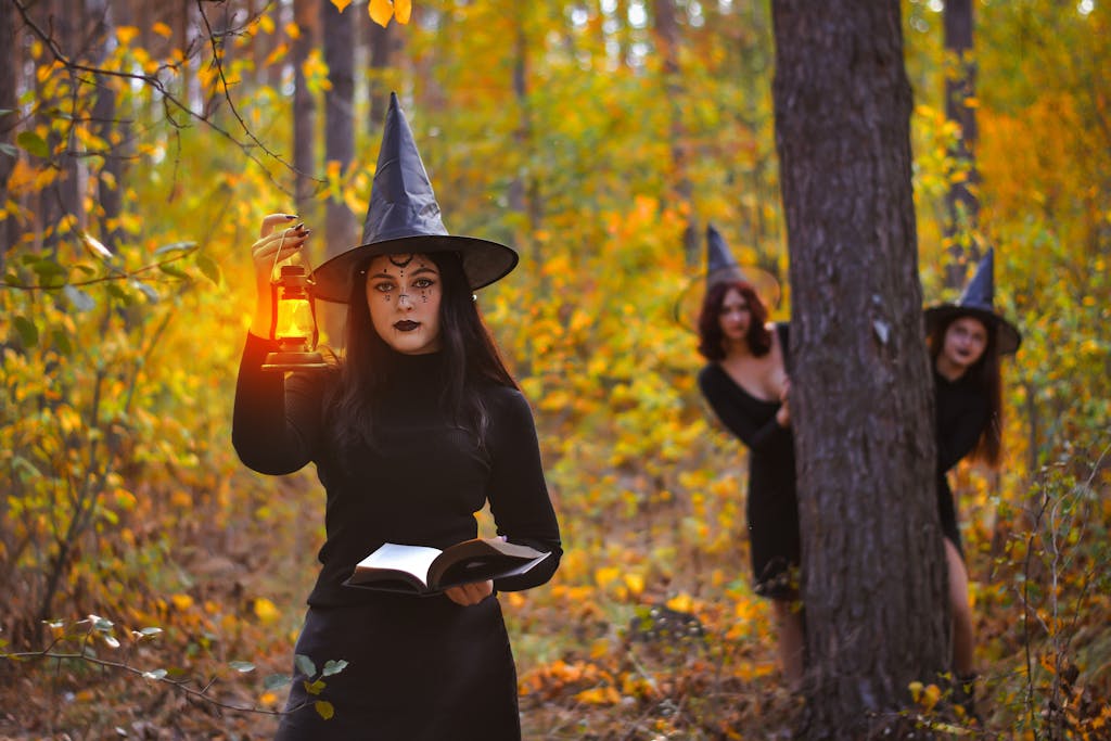 Three witches in a vibrant autumn forest, one holding a lantern, embody Halloween magic.