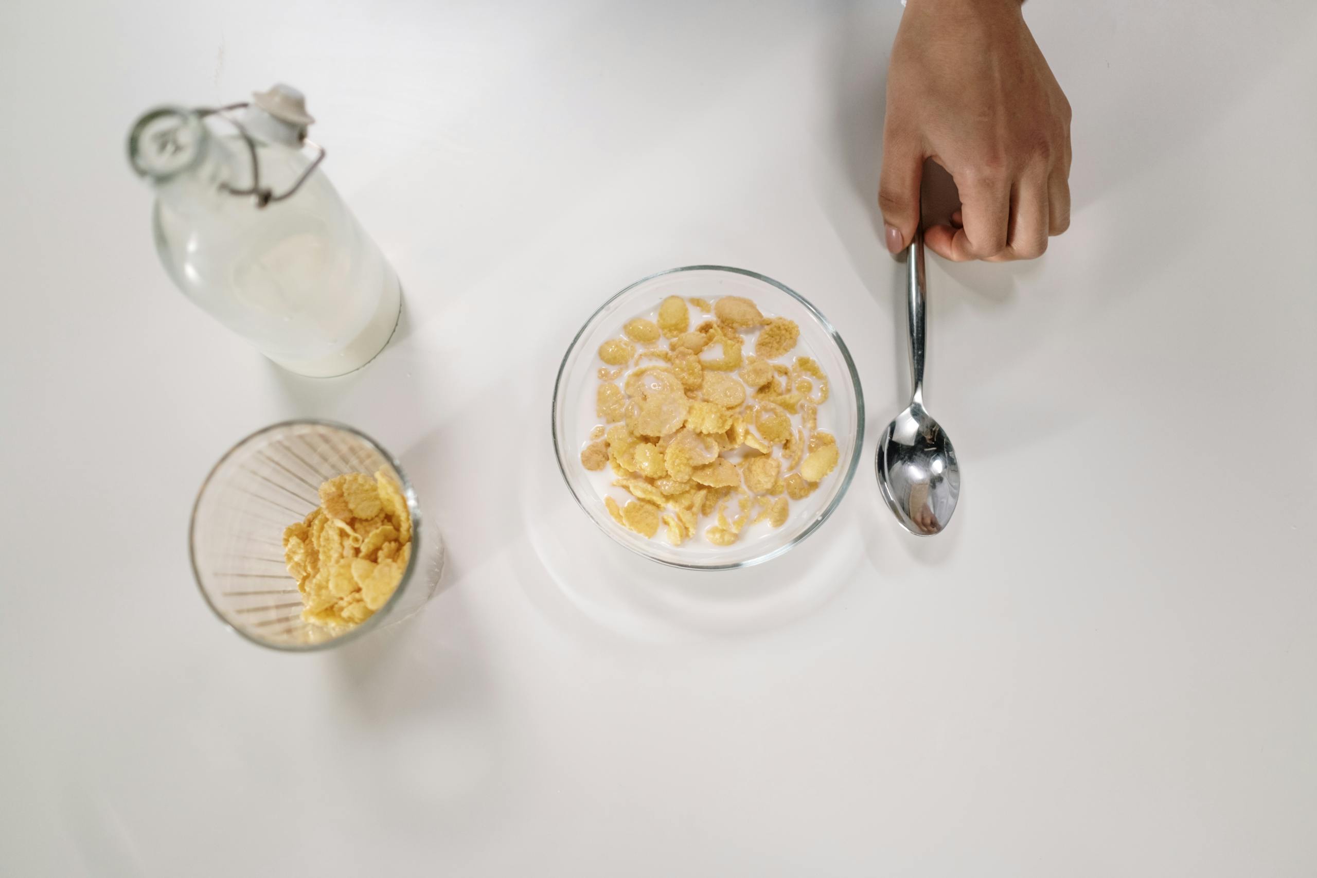Top view of a bowl of cereal with milk, a spoon, and a milk bottle on a white surface.