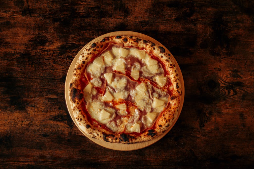 Top view of a Hawaiian pizza with pineapple and ham on a rustic wooden table.
