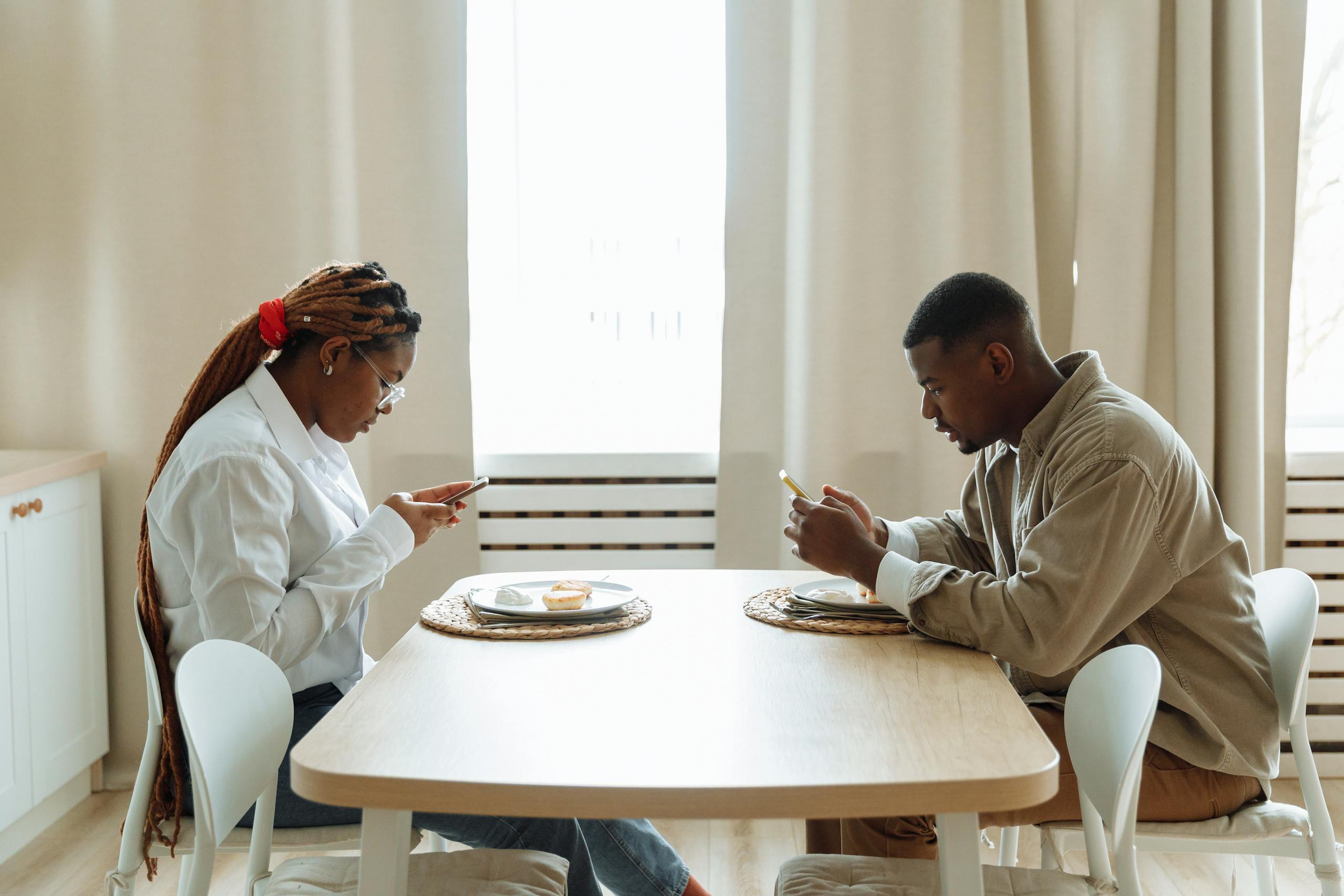 Two adults sitting at a dining table, focused on their smartphones while having breakfast indoors.