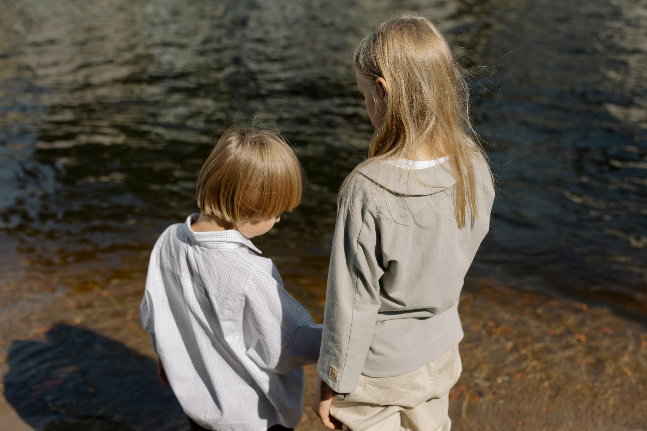 Two children stand by a river, engaged with nature, creating a serene childhood memory.