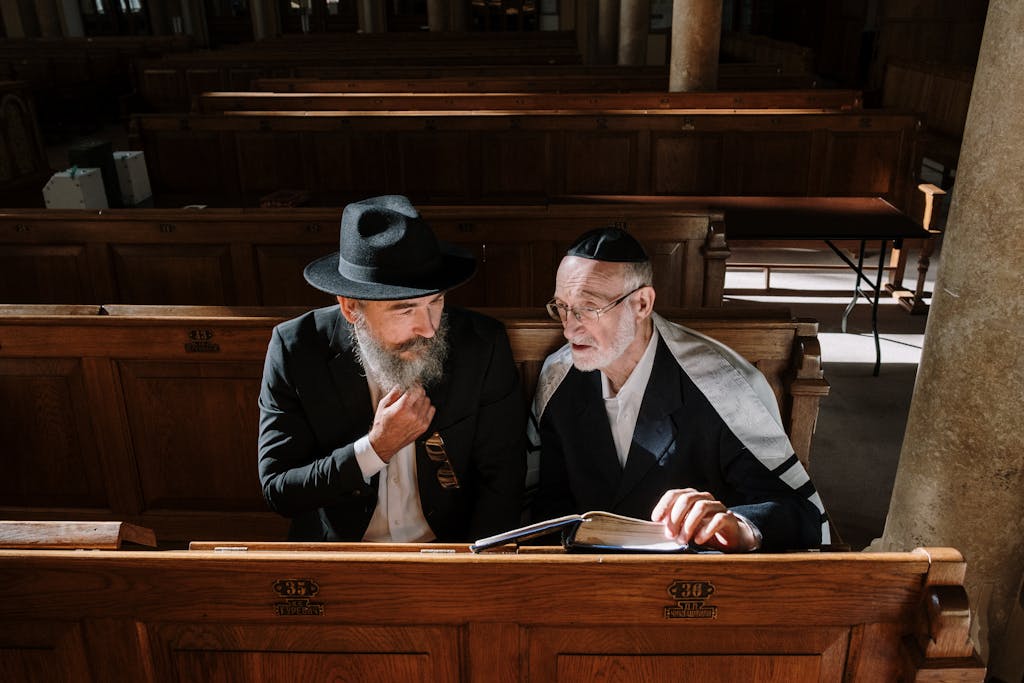 Two elderly men sitting together in a synagogue, reading a book under natural light.