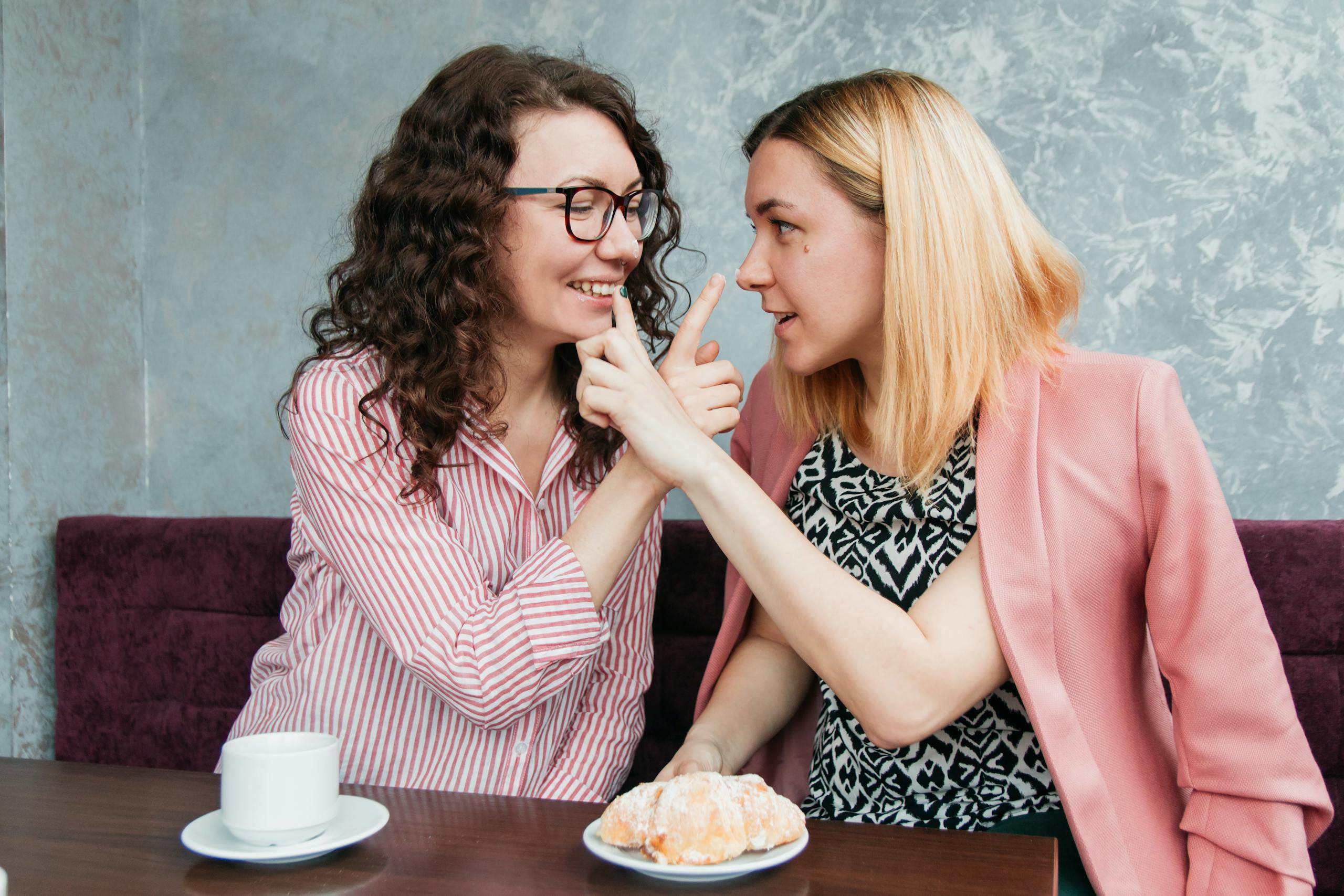 Two friends sharing a playful moment over coffee and pastry in a cozy café setting.