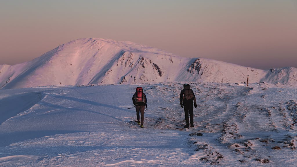 Two hikers explore a snowy mountain landscape during sunset, reflecting purple hues on the peaks.