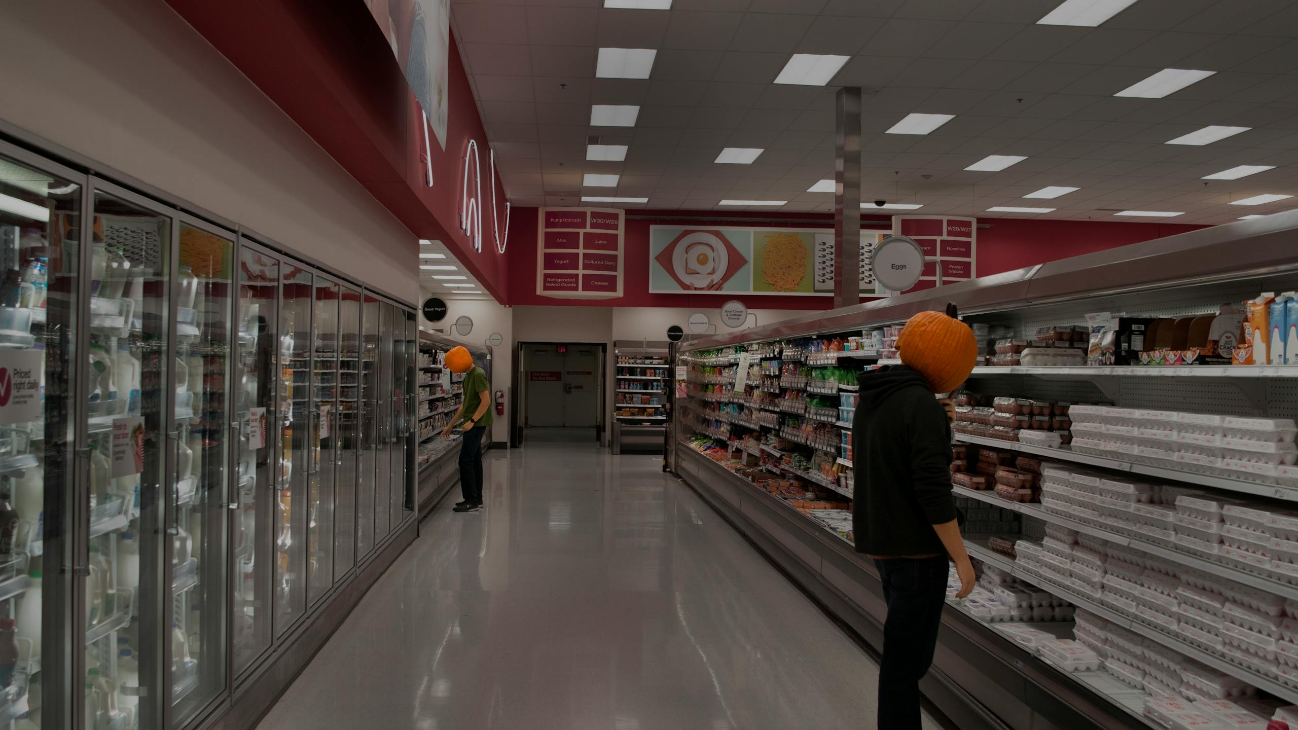 Two people wearing pumpkin masks browsing in a supermarket aisle filled with groceries.