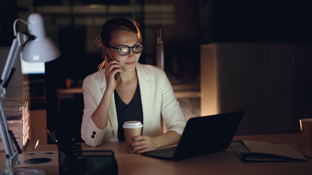 Ignore These 8 Hacks and Your Mornings Will Turn Chaotic and Stressful 1 Woman in glasses working late at night on laptop, holding coffee, making phone call.
