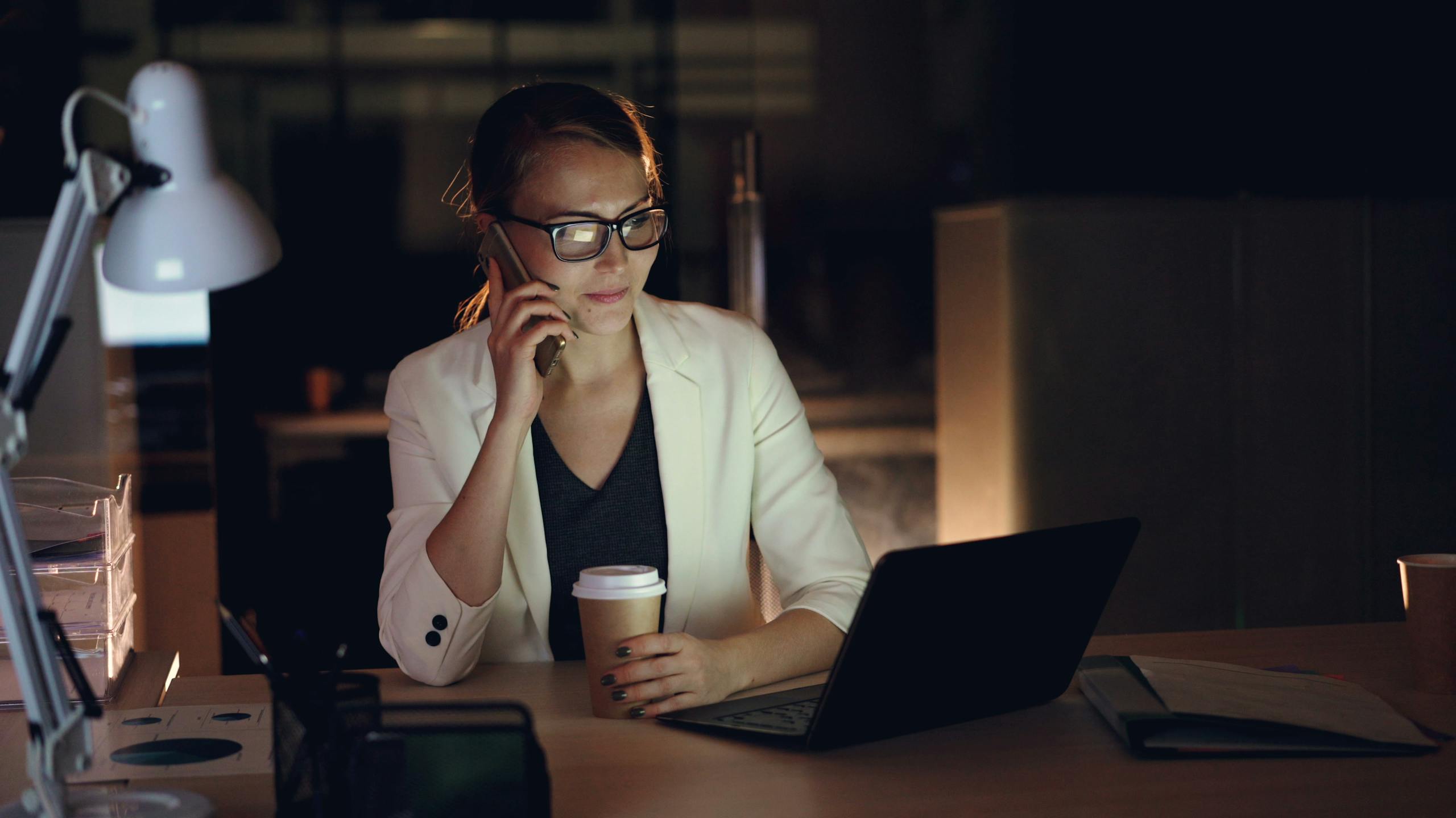 Woman in glasses working late at night on laptop, holding coffee, making phone call.