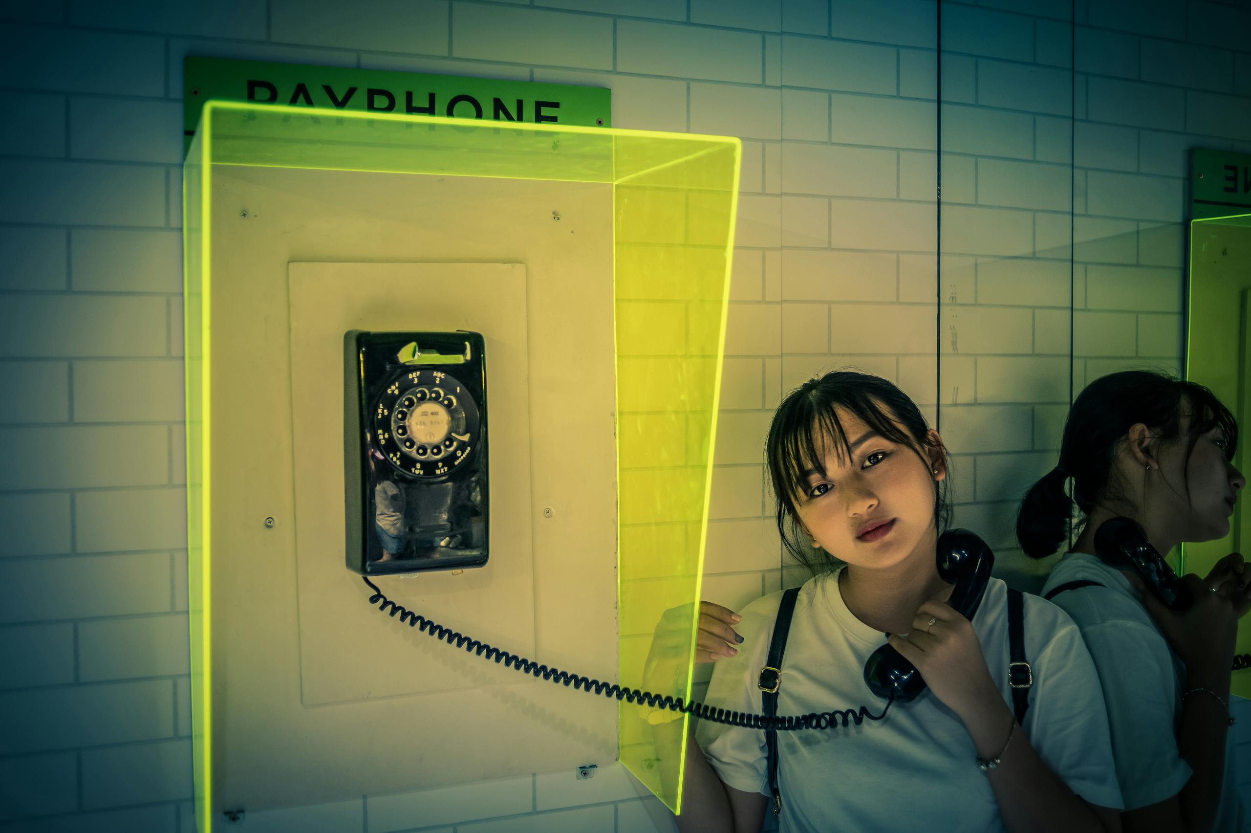 Young Asian woman posing with a vintage payphone in a stylish indoor setting. Soft light enhances the ambiance.