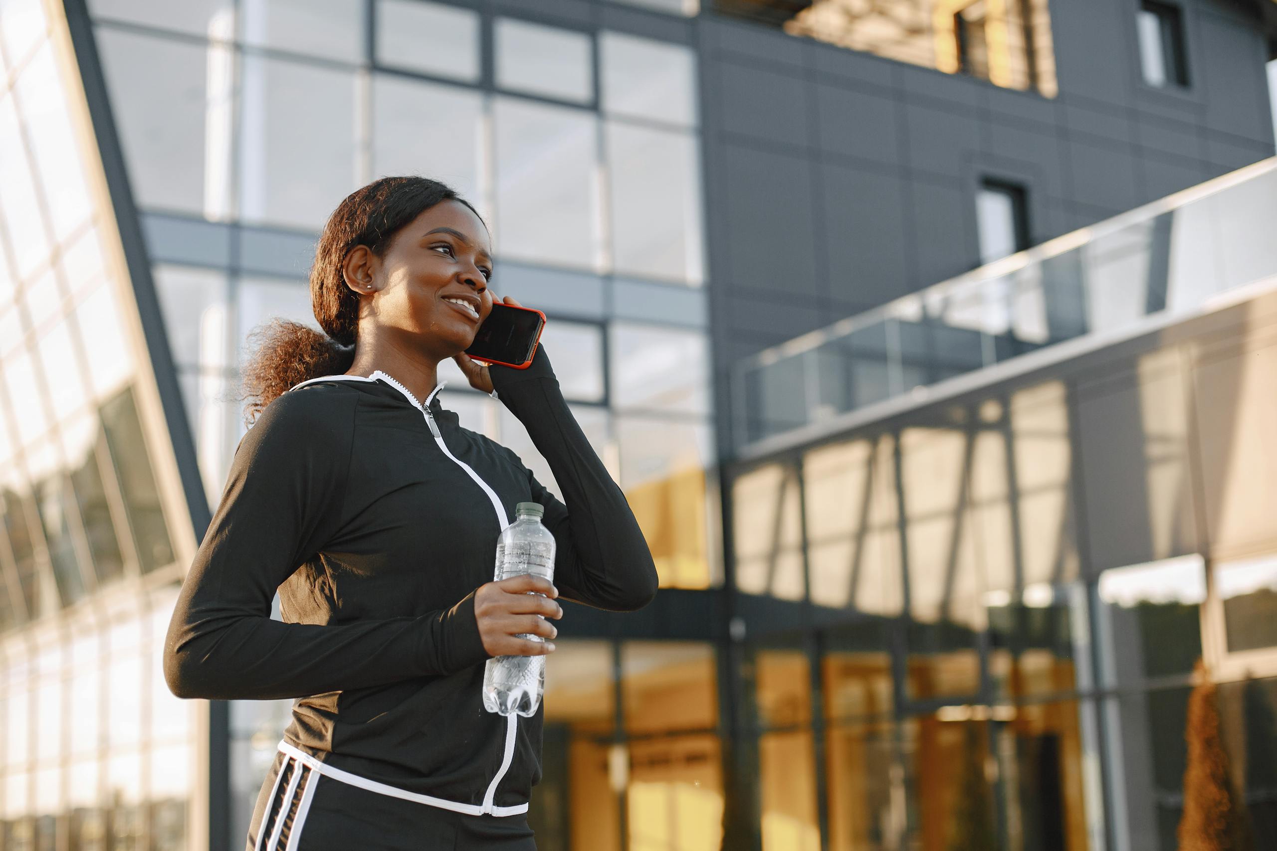 Young woman in activewear with water bottle, talking on phone outside modern glass building.