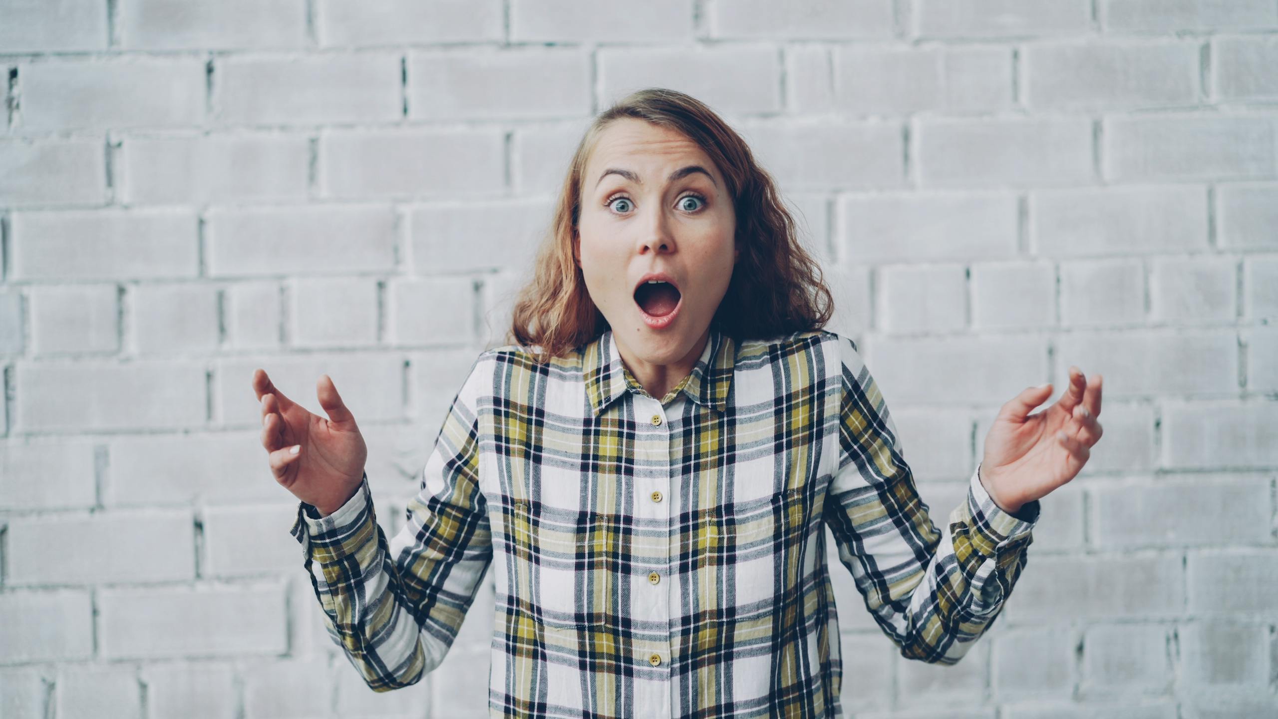 Young woman in plaid shirt expressing surprise against brick wall backdrop.