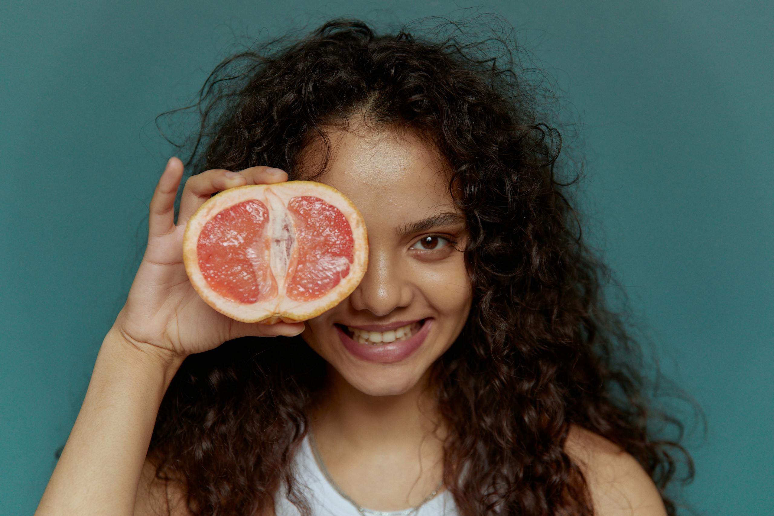 Young woman with curly hair smiling and holding a grapefruit half over her eye.