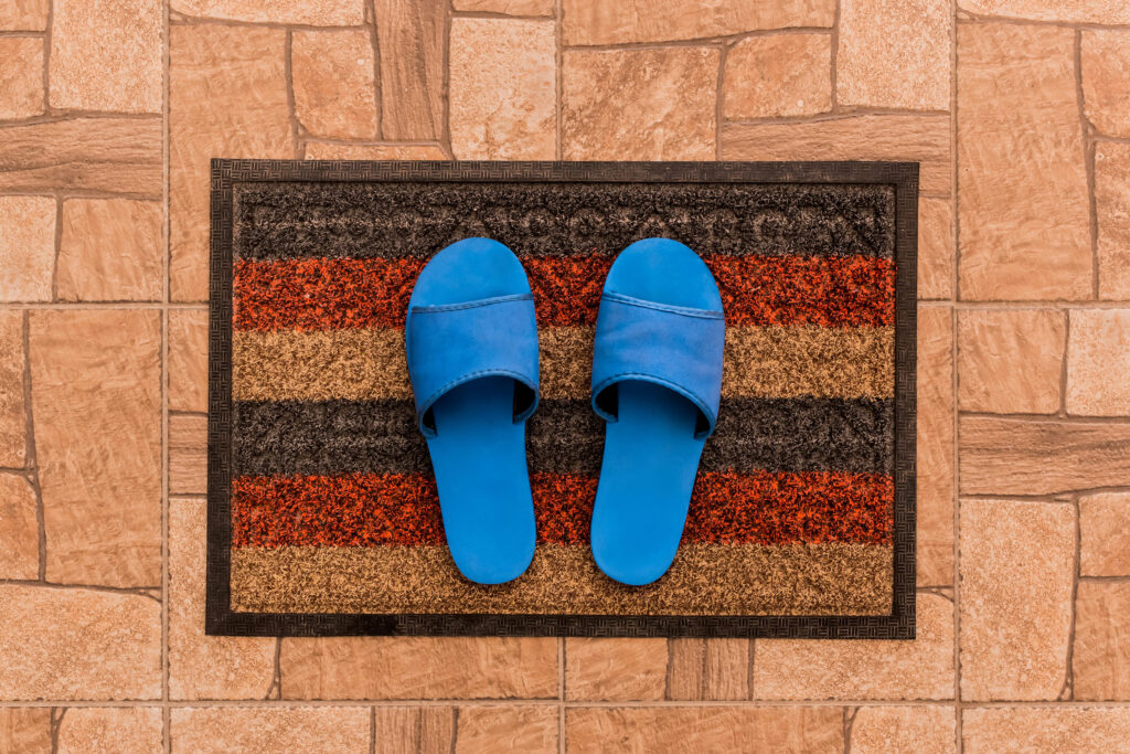 Men's blue house slippers stand on a foot mat on a brown tiled floor texture background, top view.