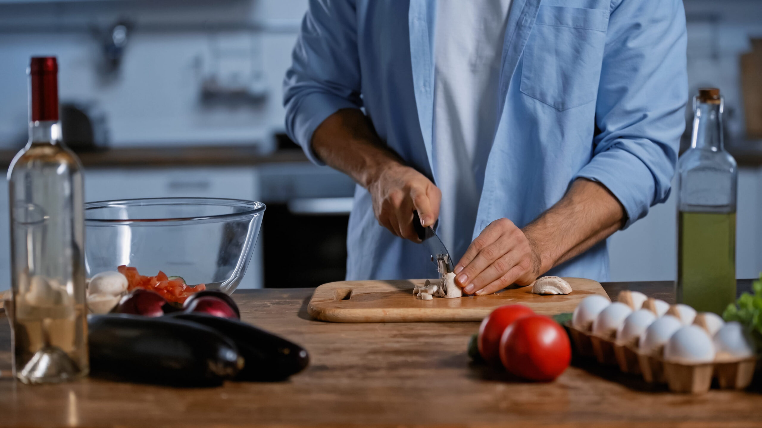cropped view of man cutting mushrooms near fresh vegetables, chicken eggs and bottle of wine