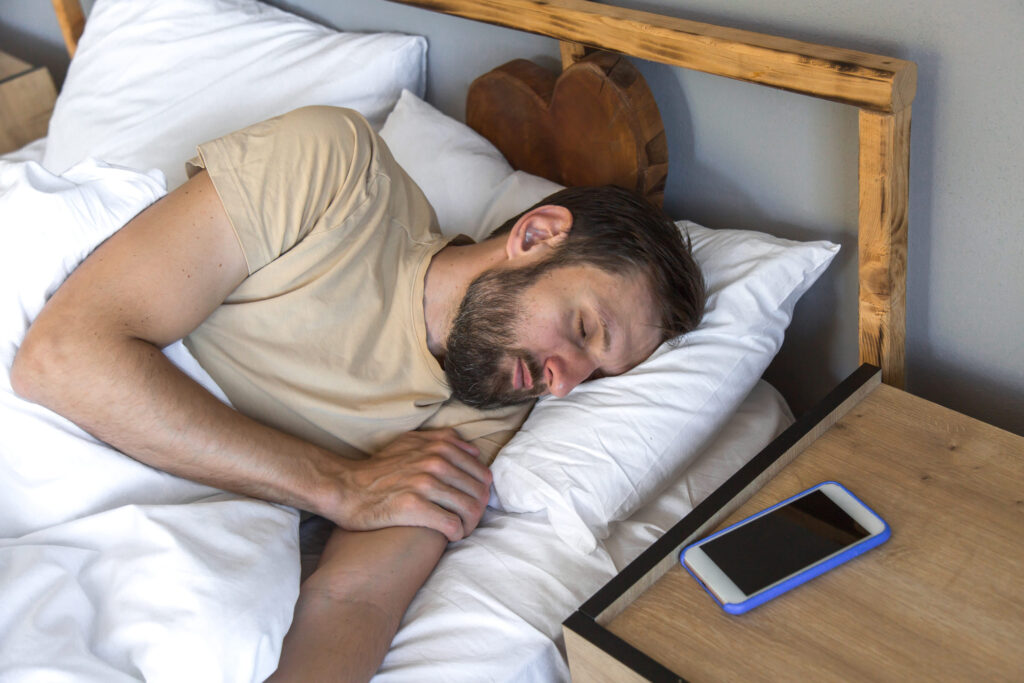 Man sleeping peacefully in bed next to smartphone on wooden nightstand, concept of digital detox and sleep quality
