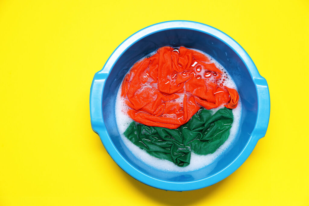 Plastic basin with clothes, water and foam on yellow background, top view