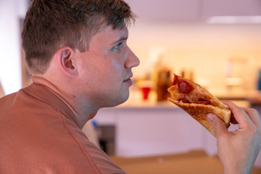 A man savors a slice of pepperoni pizza in a warmly lit indoor setting, side view emphasized.
