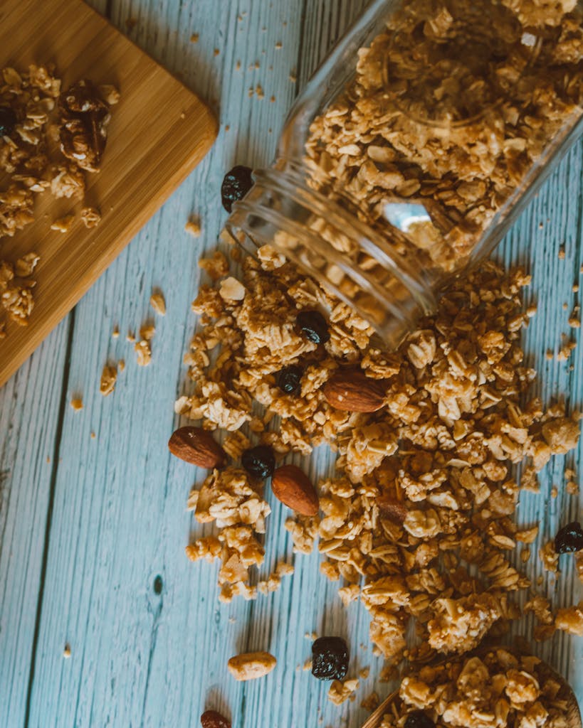 A wholesome mix of granola, almonds, and dried fruits spilled from a jar onto a wooden table.