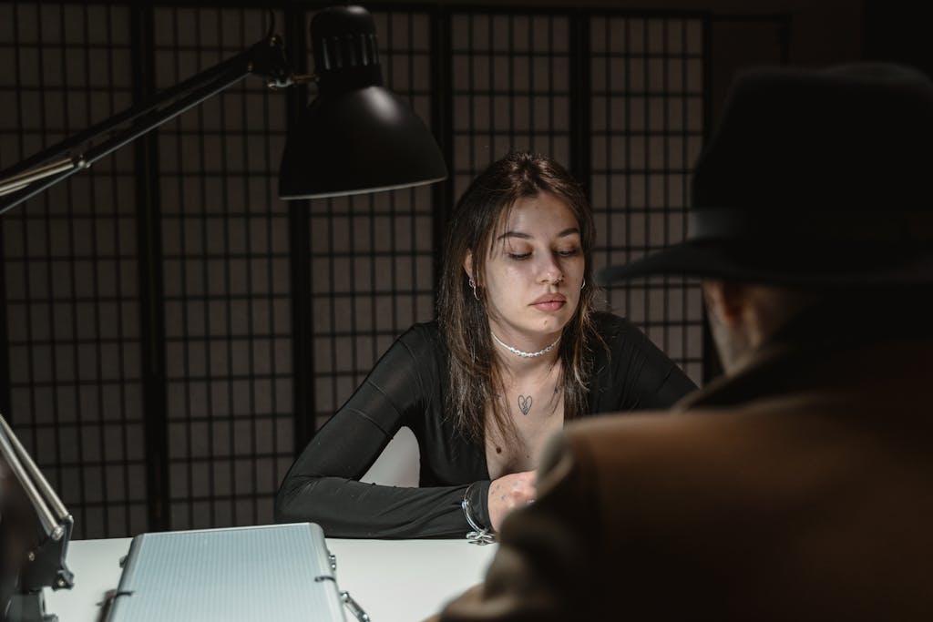 A woman and man sit across in a dimly lit interrogation room, suggesting mystery.