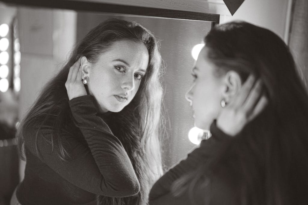A woman contemplating her reflection in a lit vanity mirror indoors.