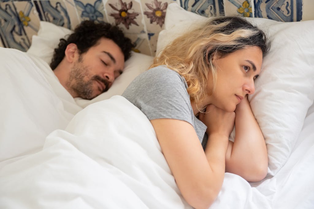 A woman lies awake next to her sleeping partner in a cozy bedroom setting, contemplating.