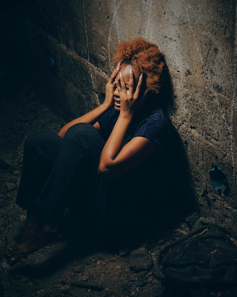 A woman with afro hairstyle sits crying against a dark wall, conveying intense emotion and sadness.