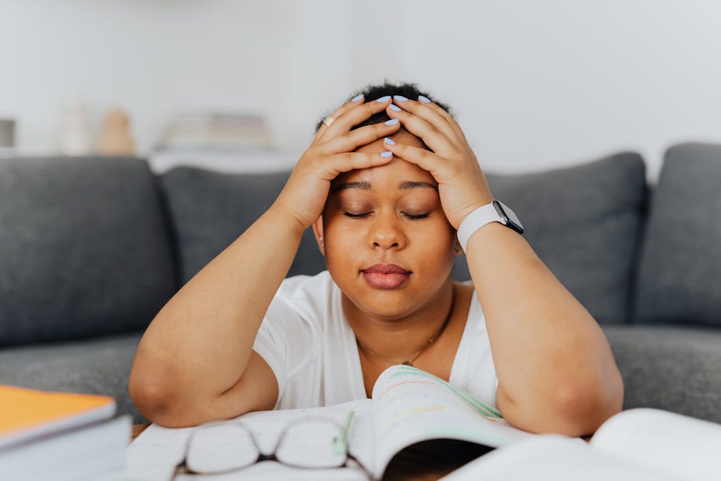 A woman with eyes closed holds her head in stress, surrounded by books in an indoor setting.